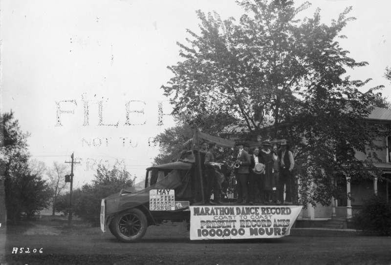 A VEISHEA parade float. People are standing on the back of a flatbed truck, two with instruments, and the float bears two large signs. One is a May calendar, mounted on the truck door. The other sign, centered on the truck bed, reads:"Marathon Dance Record / Coast to Coast / Present Record Ames / 1000,000 Hours.".