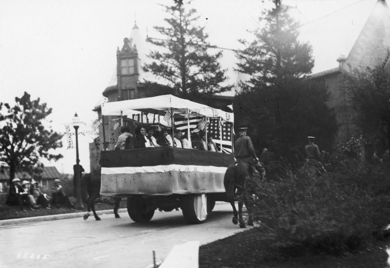 1923 motorized vehicle float with three member of ROTC in the front seat. Seated on the flat bed of the float are nine women, with another ROTC member behind them. ROTC members on horseback ride along side the float. Margaret Hall can be seen in the background. See photograph 1722-5-6 for an alternate view.