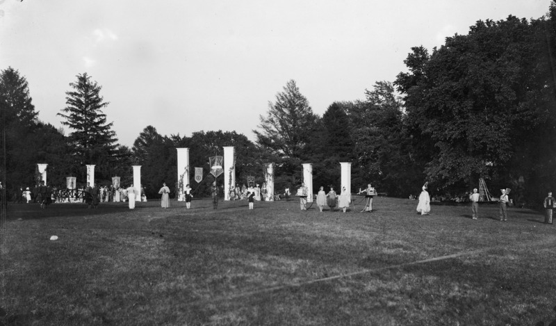 A 1920s central lawn event that may or may not be associated with VEISHEA. A number of people are present in the midst of eight simulated columns on the lawn. People are in costume, some holding exhibit like materials, and others holding banners.