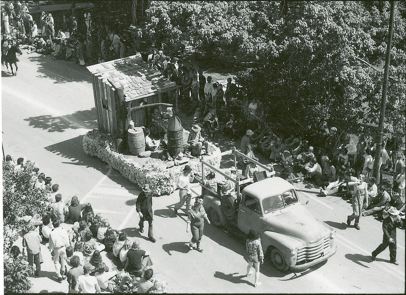 A 1976 VEISHEA parade float. An old model pickup truck tows a float depicting a hillbilly scene with a shed and a still. Several participants are dressed in overalls.