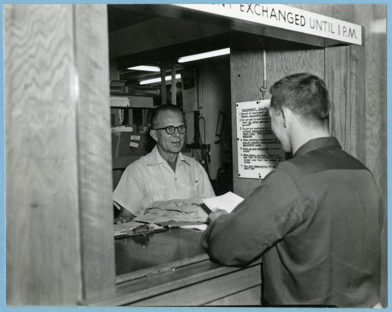 Orville Hagen, behind the counter, issues equipment of Mack Jerkatis, freshman wrestler, 1956.