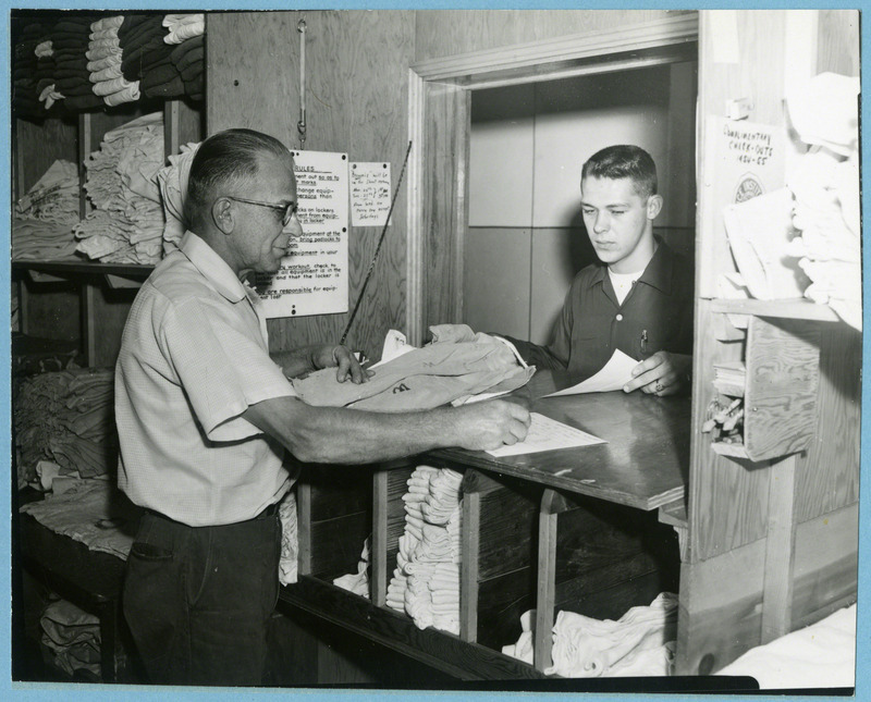 Orville Hagen, behind the counter, issues equipment of Mack Jerkatis, freshman wrestler, 1956.