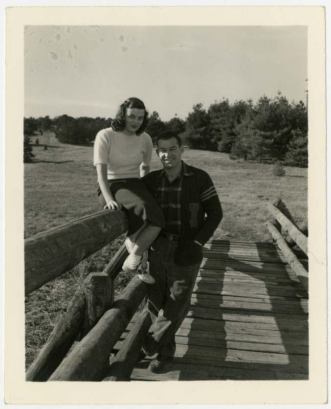 Glenn Brand, Iowa State University wrestler, is standing on a wooden bridge. A girl is sitting on the fence beside him, 1946.