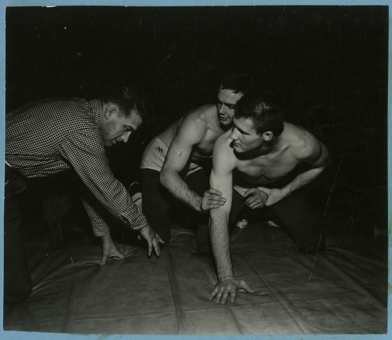 Two unidentified men practice grappling. Coach Harold Nichols coaches from the side.