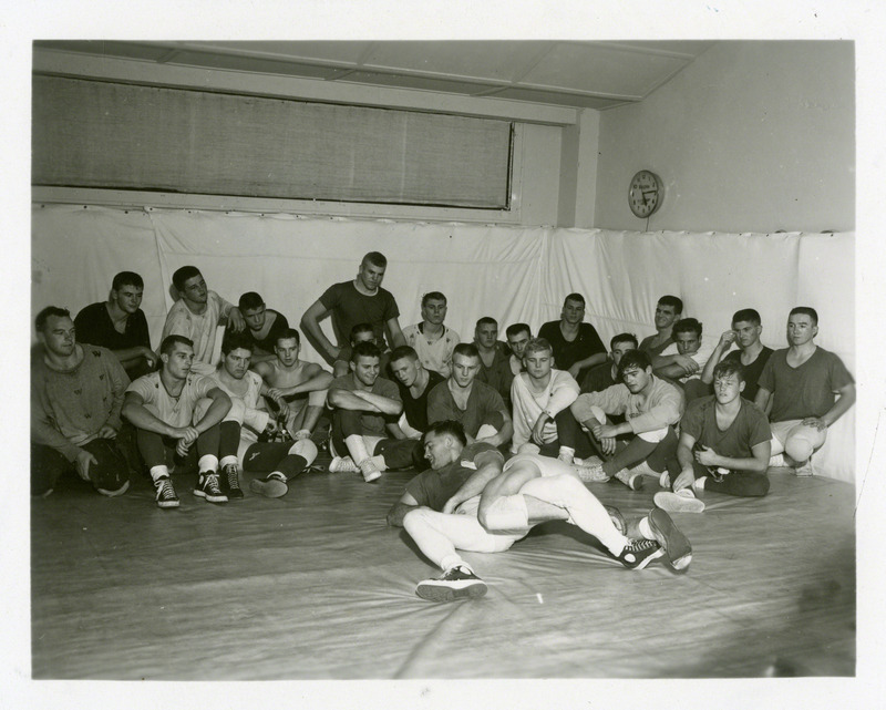 Two unidentified men practice grappling as a group of men watch in the background.