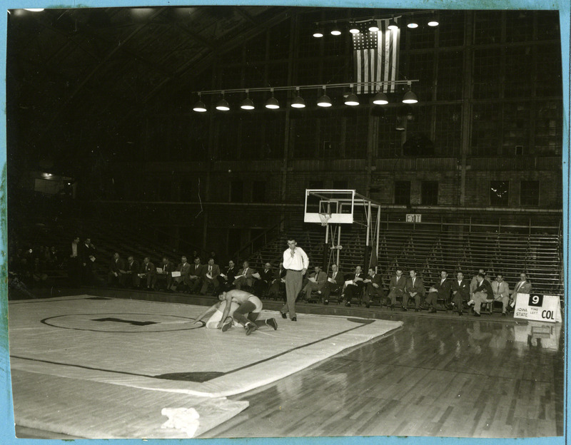 Two unidentified men grapple at a reunion of wrestling lettermen. A line of wrestling lettermen can be seen in the background.