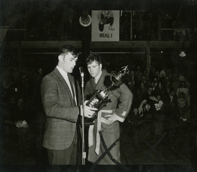 An unidentified man, holding a trophy, speaks to Dan Gable.