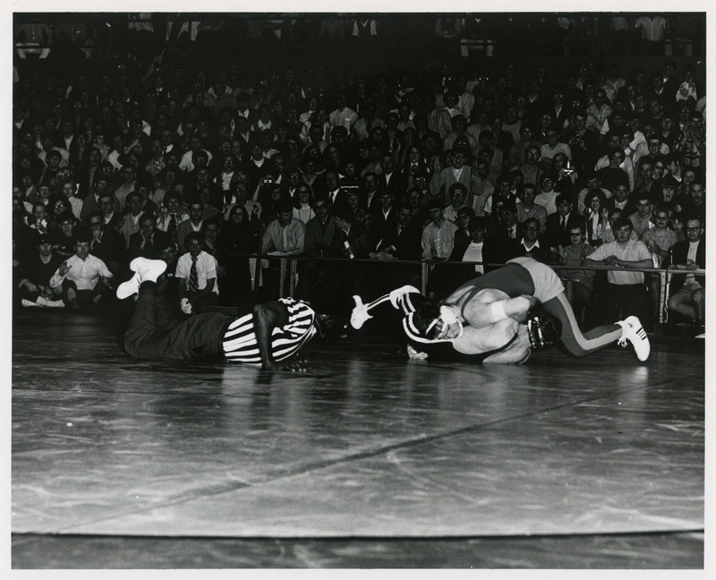 Two men grappling at wrestling event. Referee is on the ground watching.