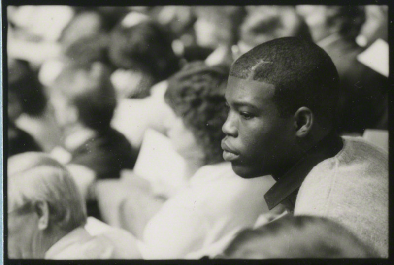 Close-up of a student at the memorial service. Audience members are in the background.