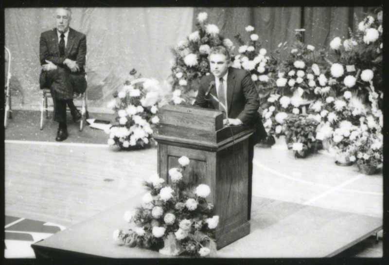 Max F. Urick, Director of Intercollegiate Athletics, is speaking from the podium at the memorial service.