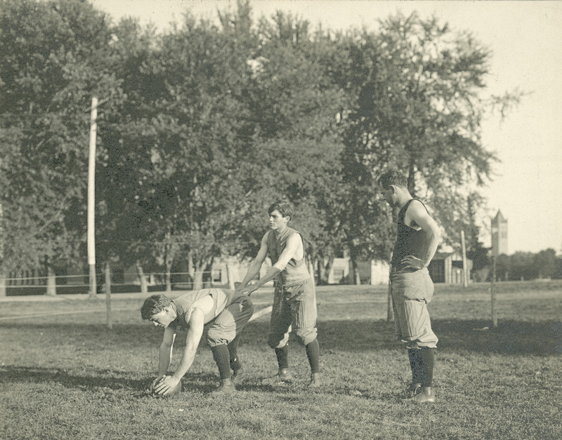 Three individuals playing football. The Campanile and other buildings can be seen in the background.