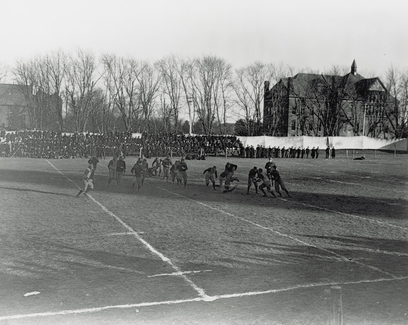 A football game in progress on the first athletic field. Spectators view from the stands. Morrill Hall can be seen in the background on the right side of the image.