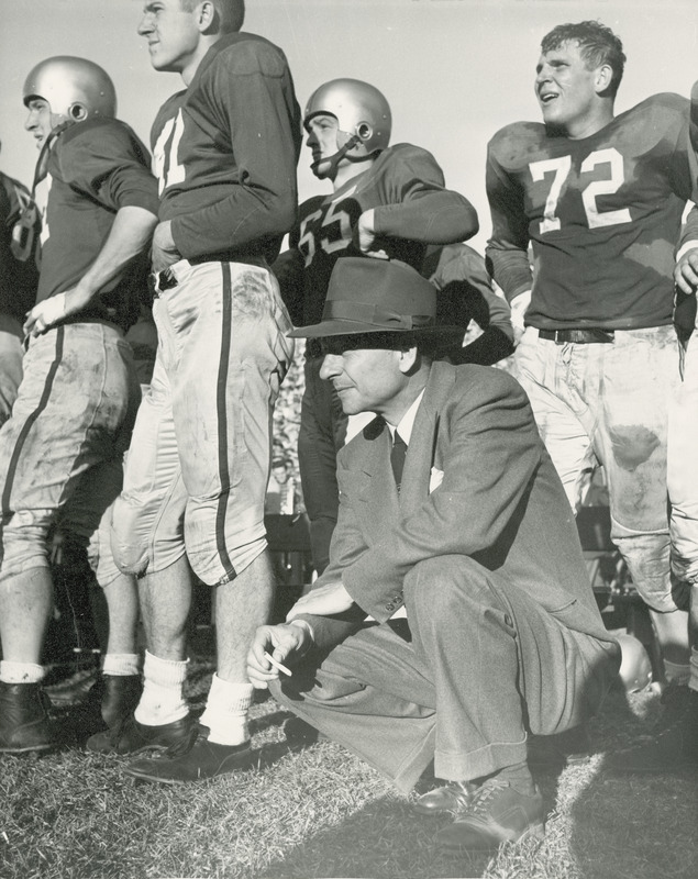 Coach Emmet R. "Abe" Stuber squatting. Coach Stuber is wearing a suit and hat. He is surrounded by standing football players in uniform.