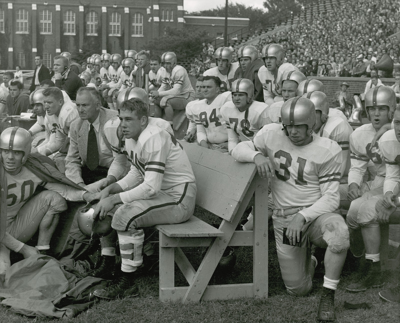 The sidelines during a football game. The football players and coaches are in the foreground, while spectators are in the background. State Gymnasium can also be partially seen.