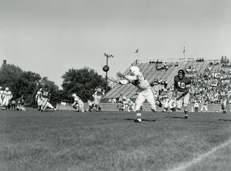 A football game in progress before a partially filled stand of spectators. Football player in motion to receive the ball.