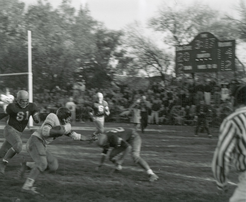 A football game in progress. The football players are in the foreground, spectators and scoreboard are in the background. Scoreboard indicates Iowa State 20, visitors 14.