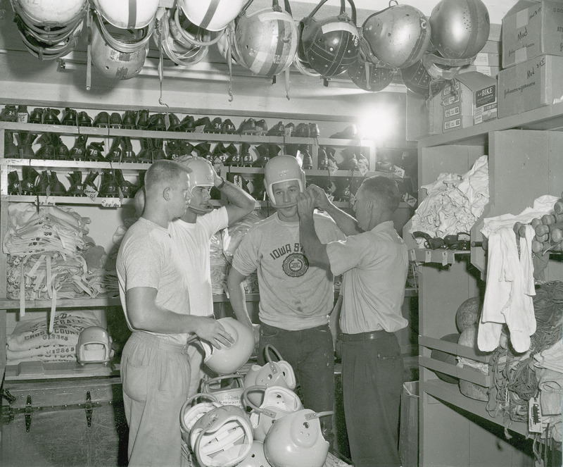 Three males in an equipment room trying on football helmets. A fourth male is assisting them.