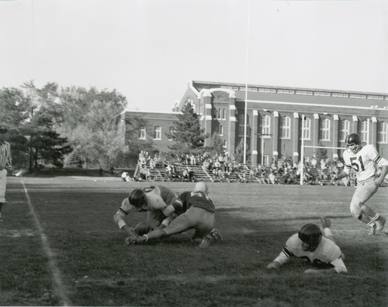 A football game being played before a group of spectators. State Gymnasium is in the background.