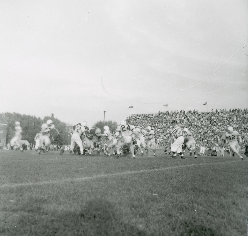 A football game in progress. The players and a referee are in the mid-ground and the spectators are in the background. This image is part of a set of images that includes: 2-46.G.Football.1871-5-6-1.