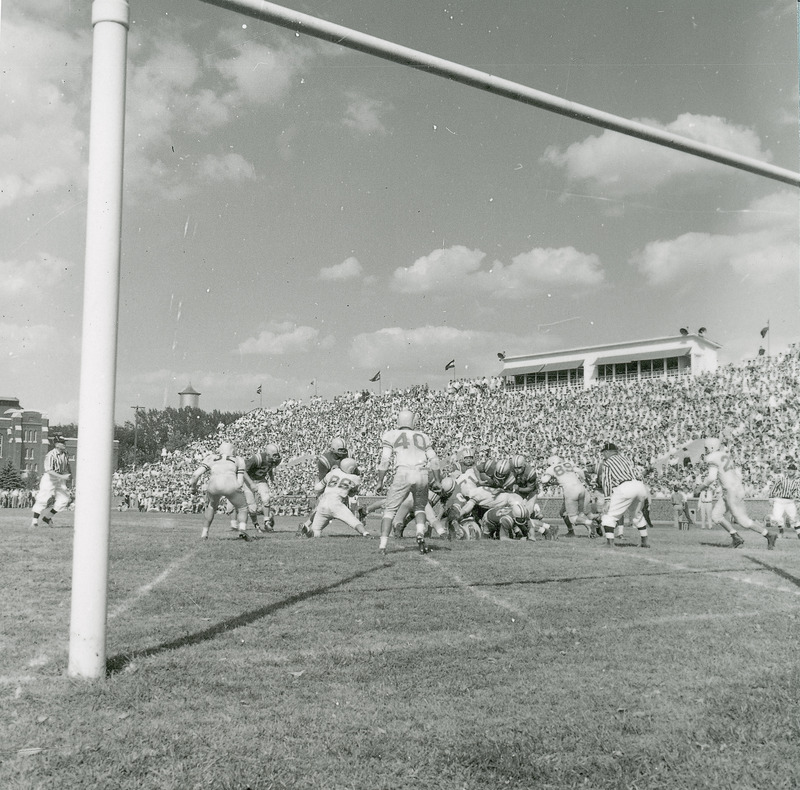 A homecoming football game in progress. There is a stadium full of spectators in the background, the players in the mid-ground and the goalposts in the foreground. In the distant background, the top of the Marston Water Tower is visible over the trees. This image is part of a set of images that includes: 24-6-G.Football.1871-5-7-1; 24-6-G.Football.1871-5-7-2 ; and 24-6-G.Football.1871-5-7-4.