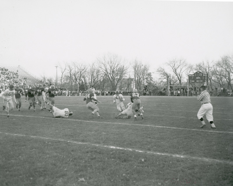 A football game in progress. The football players and a referee are in the foreground. Spectators are seated in the bleachers and stand outside the end zone.