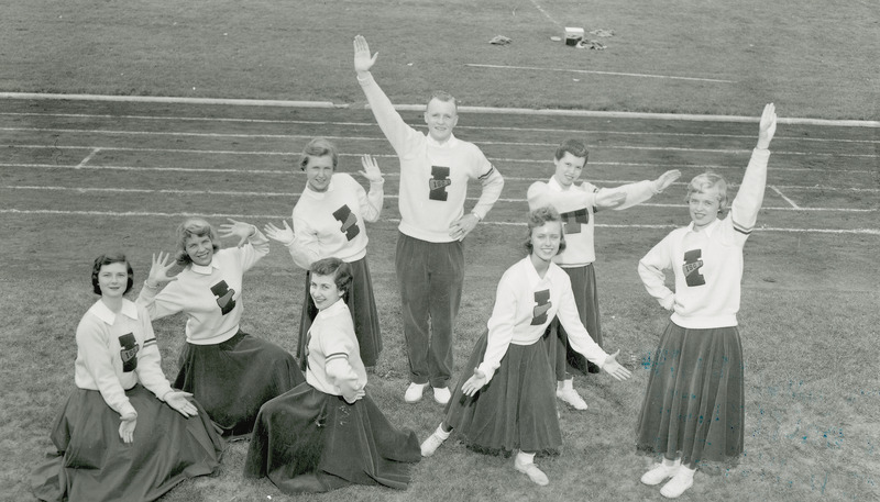 Seven women and one man are posed in their cheerleading uniforms. Maxi skirts and long sleeve sweaters with shirt collars for the women, and long pants and the same sweater for the gentleman.