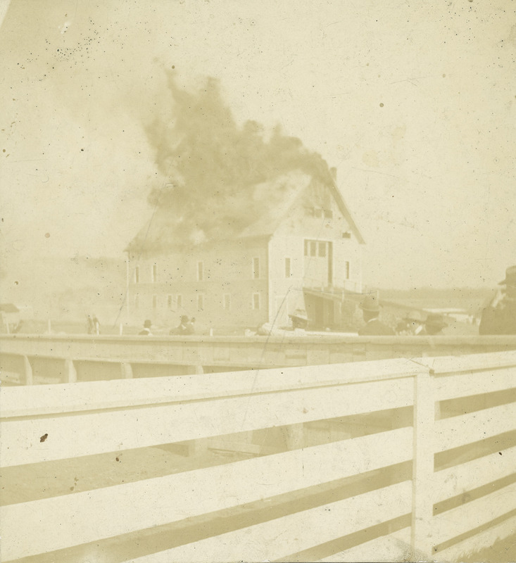 The fire at the Iowa Agricultural College Experiment Barn in 1902. The building was of frame construction with a gable roof. Flames are engulfing the roof as a number of onlookers watch.