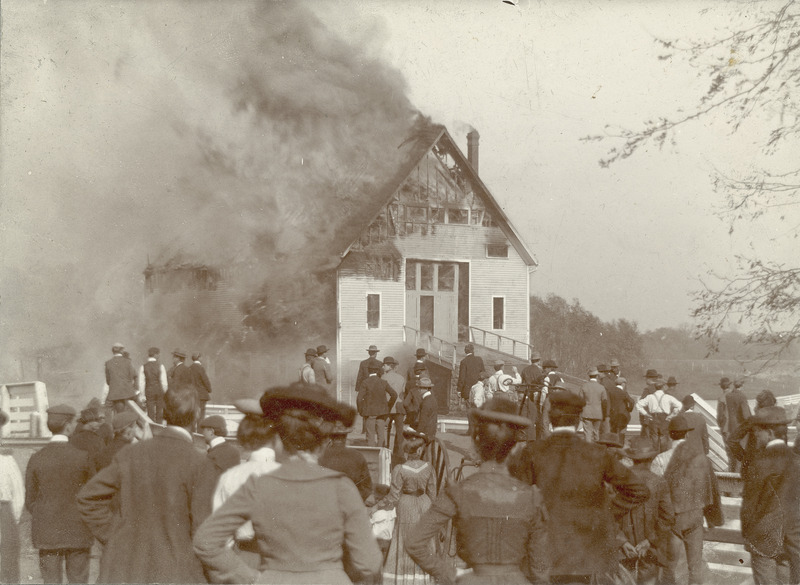 The fire at the Iowa Agricultural College Experiment Barn in 1902. The building was of frame construction with a gable roof. Flames are engulfing the roof and traveling down the sides of the building as a crowd of onlookers watches.