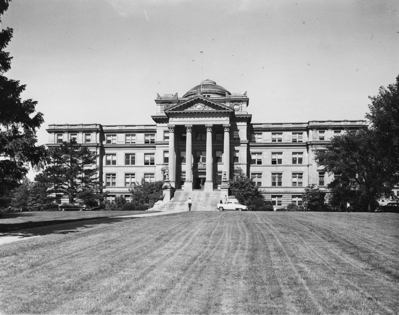 The front of Beardshear Hall. Vehicles are parked on the street and four people are on the sidewalk and steps.