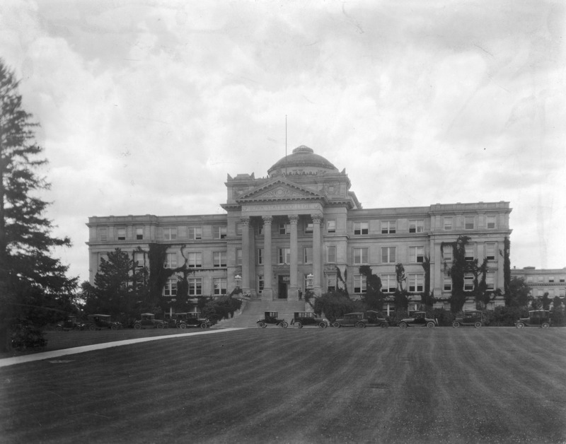 The front of Beardshear Hall. 1920s vehicles are parked on the street in front of the building.