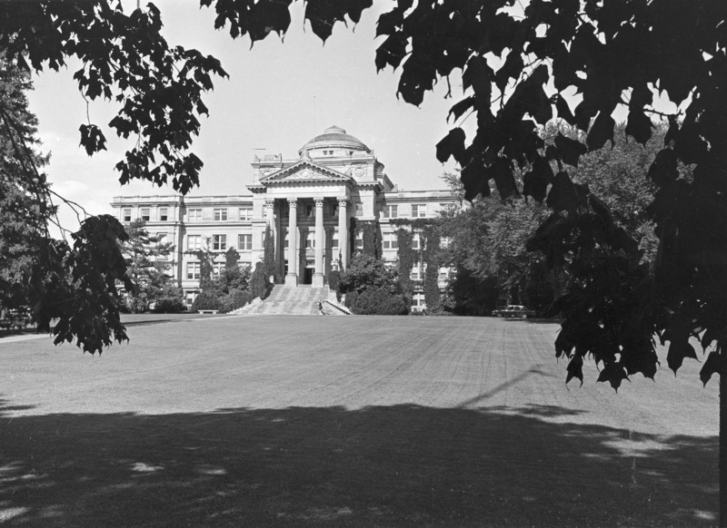 The front of Beardshear Hall. The image is framed by overhanging tree limbs in leaf. A single car is parked in front of the building; two people are on the sidewalk in front of the building.