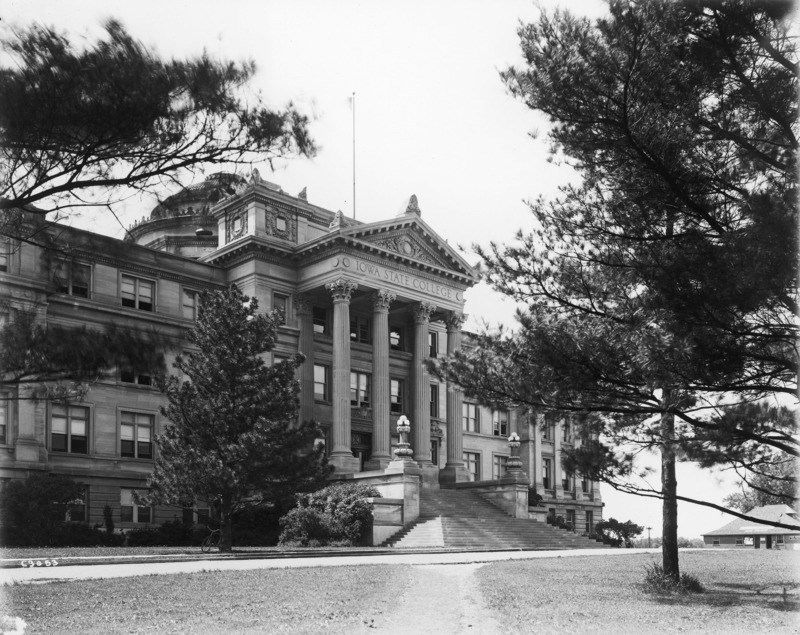 The front of Beardshear Hall, with a dirt walkway angling toward the bottom of the steps.The Hub (Dinkey Station) is in the distance on the right of the image. A bicycle is leaning against a tree.