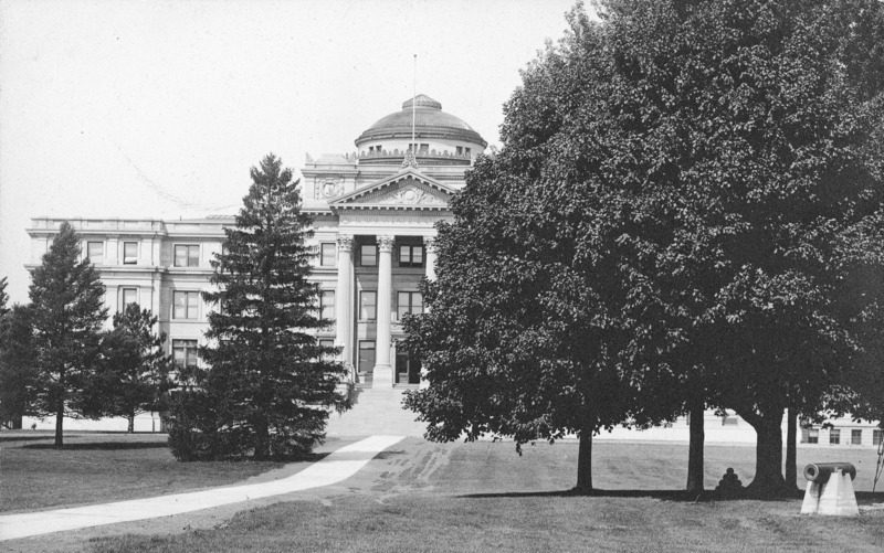 The front of Beardshear Hall with a tree grove and cannon on the central campus lawn obscuring the view of the right side of the building.