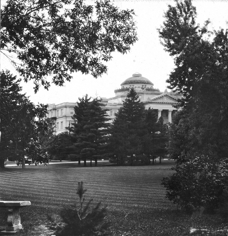 The roofline of Beardshear Halland part of the portico. The rest of the building obscured by trees on central campus. A stone bench is in the foreground. A single car is parked on the street.