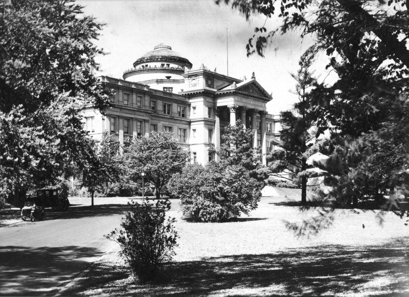 The front of Beardshear Hall from an angle. The building is surrounded by numerous trees and shrubs. A 1920s vehicle and a motorcycle with a side car are parked on the street.