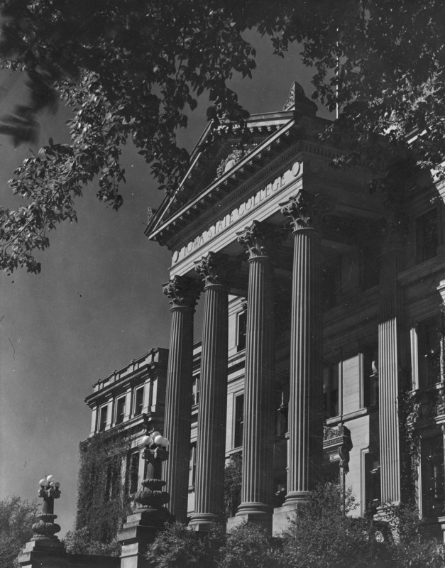 The Beardshear Hall front portico, emphasizing the entablature, the Corinthian columns, and the Beaux Arts step lamps. The image is framed by overhanging tree limbs.