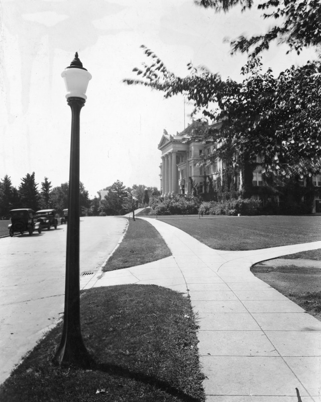 The front of Beardshear Hall from an angle. A street lamp is prominent in the foreground. 1920s vehicles are parked on the street. Ivy vines are growing on Beardshear Hall, which is partially obscured by a tree limbs on the image right.