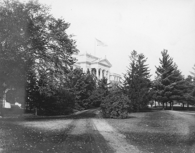 The front of Beardshear Hall much obscured by trees and shrubs.A dirt road leads toward the Beardshear Hall front steps. The English Office Building is seen through the otherwise obscuring tree foliage on the left of the image. A flag is flying from the flag pole on top of Beardshear Hall.