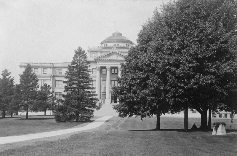 The front of Beardshear Hall as seen through trees that obscure the view of the left and right of the front of the building. Central campus trees and a cannon are on the right side of the image.