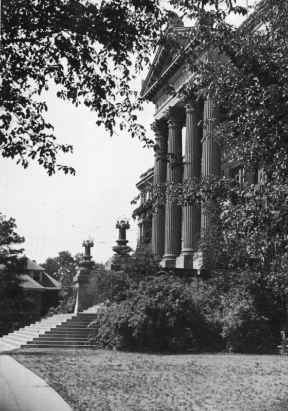 The portico and front steps of Beardshear Hall as seen from the north. Old English Office building is in the image in the background. Shrubs close to the steps and overhanging fully leaved trees frame the image.