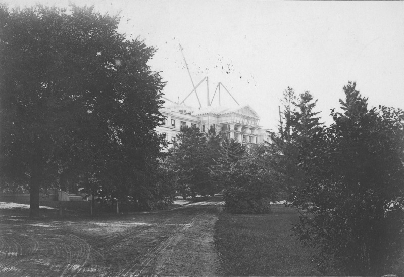 The front of Beardshear Hall when construction is nearly complete. Scaffolding is on the front columns and lifting rigging is on the roof.The view of the building is largely obscured by trees and shrubbery. A dirt road leads toward the front of Beardshear through the trees.The front steps of English Office Building can be seen through the obscuring trees on the left of the image.