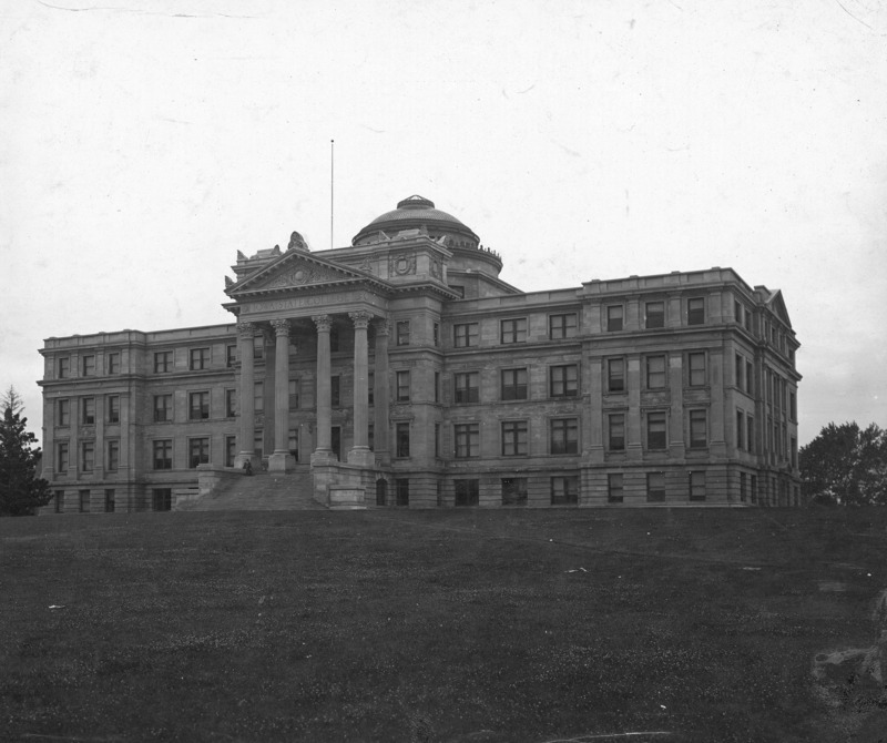 The front of Beardshear Hall without any landscaping material in front of the building. The clover filled lawn in front shows student paths but no formal walks or streets. A person is sitting on the plinth at the top of the steps.