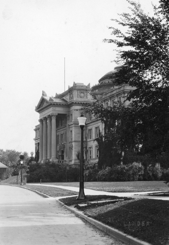 The front of Beardshear Hall from an angle, with lush landscaping material in front of the building. The back portion of English Office Building can be seen on the left of the image. Streetlamps are located along the curb.