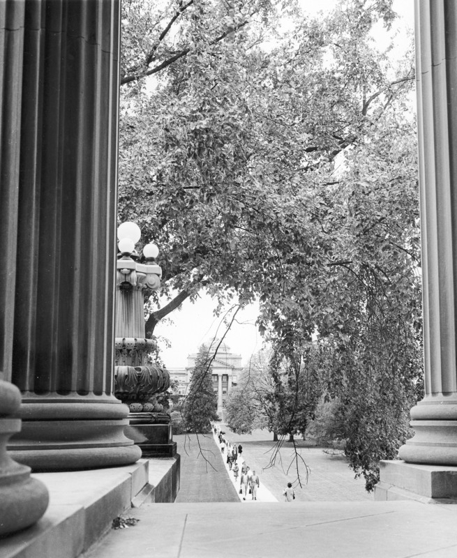 The front of Beardshear Hall seen from under the portico of Curtiss Hall across central campus. Framed by the Beaux Art details of columns and lamp post along with an overhanging mature elm, the view shows students on the central walkway.