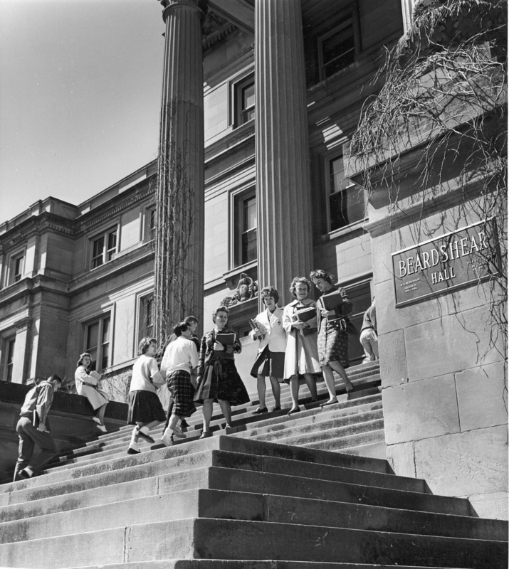 The front steps of Beardshear Hall in a close up. Details of columns and stair abutments frame ten students going and coming. A "Beardshear Hall" building name plaque is shown mounted to a stair abutment.