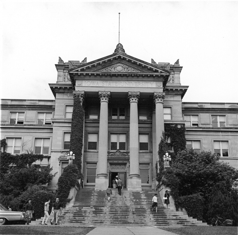 The front steps and portico of Beardshear Hall, framed by lush mature landscape material and vines on columns and building. Ten people are in the photograph. A bicycle with a wire basket stands on the sidewalk. The rear of an automobile is seen parked at the left of the image.