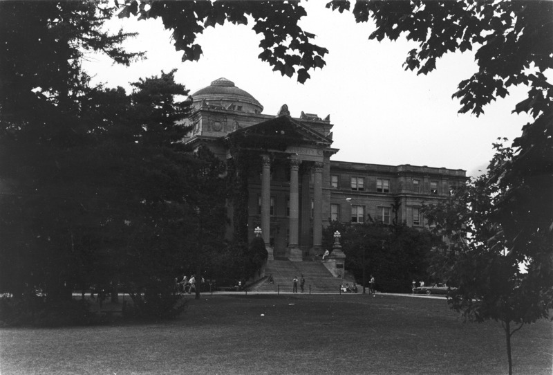 The front of Beardshear Hall, framed by dark mature foliage. Students are on the sidewalk as well as sitting on the main steps. A car is parked on the street.