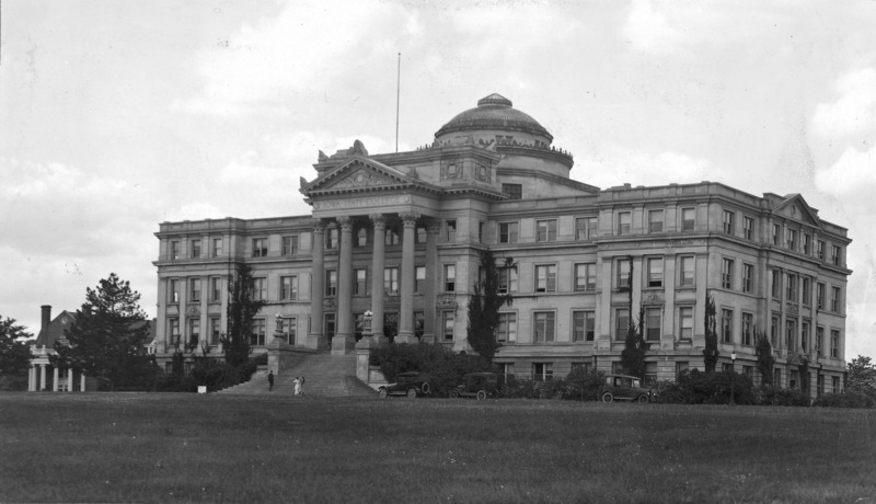 The front of Beardshear Hall.Three 1920s vehicles are parked on the street in front of the building. Three people are in front of the building.