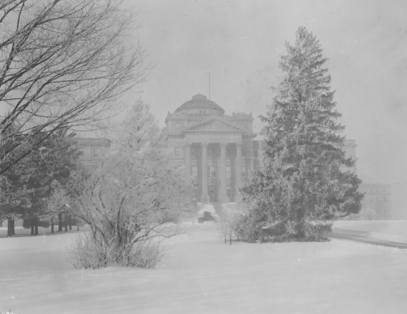 The front of Beardshear Hall from central campus. Trees and shrubs are covered in snow. 1920s vehicles are parked on the street.