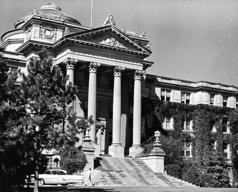 The front of Beardshear Hall from a near distance, emphasizing the portico, steps and dome. The photograph shows one person walking in front of the building.Two bicycles and one car are parked in front of Beardshear on either side of the steps.